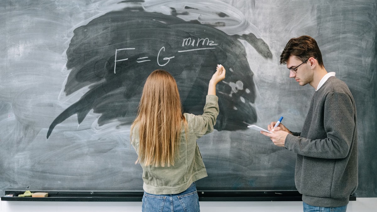 Two students discuss and solve a physics equation on the chalkboard.